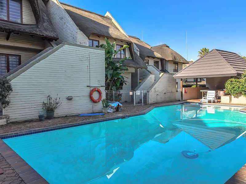 A communal swimming pool area featuring a thatched roof structure, brick paving, and perimeter fencing, set against the exterior facade of the apartment complex.