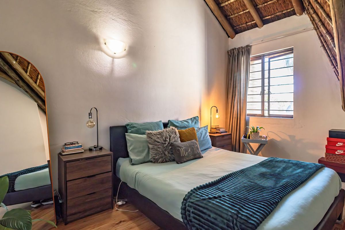 A cozy bedroom featuring a high vaulted ceiling with exposed thatch and timber rafters, laminate flooring, and a louvered window for natural light.