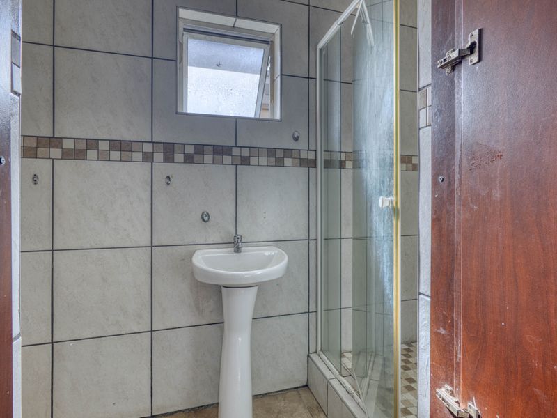A compact bathroom featuring a pedestal basin, a shower enclosure, and tiled wall finishes, requiring some maintenance and cosmetic updates.