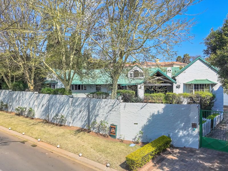 A street-facing view of a residential property featuring a painted perimeter wall, metal gate, and a pitched metal roof set behind mature trees.