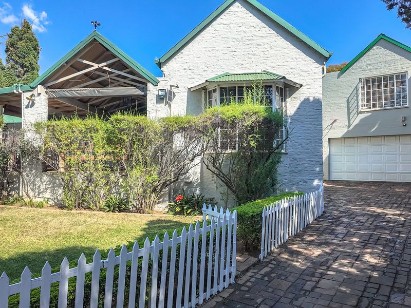 Charming front elevation featuring a paved driveway, double garage, white picket fence, and a covered patio area with exposed timber roof trusses.