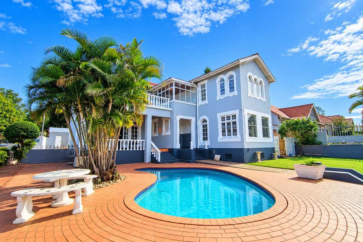 Rear exterior view of a two-story house featuring a swimming pool, brick paved patio area, and a balcony overlooking the garden.