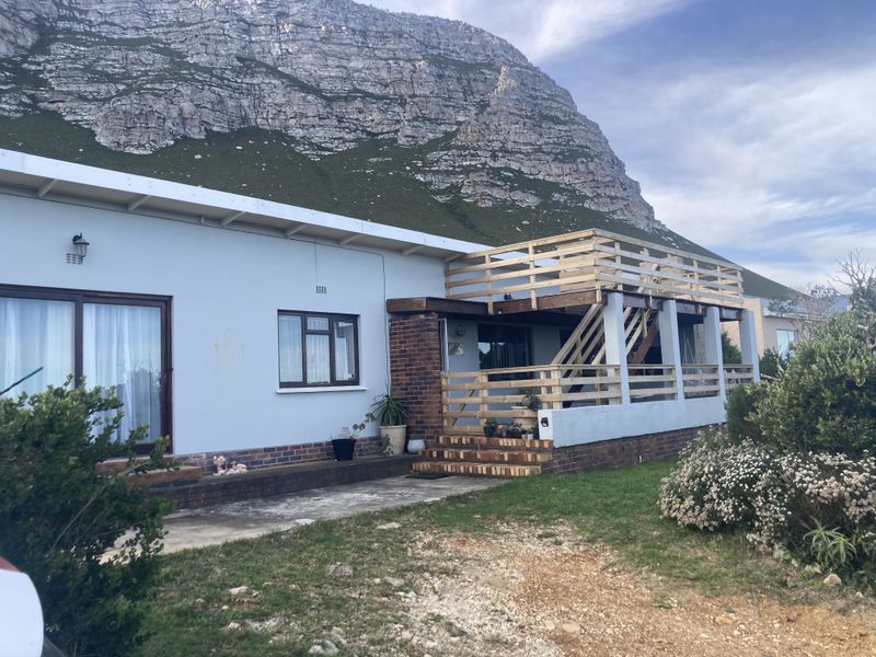 Rear exterior view of the property featuring a timber deck, staircase, and concrete patio set against a mountain backdrop.