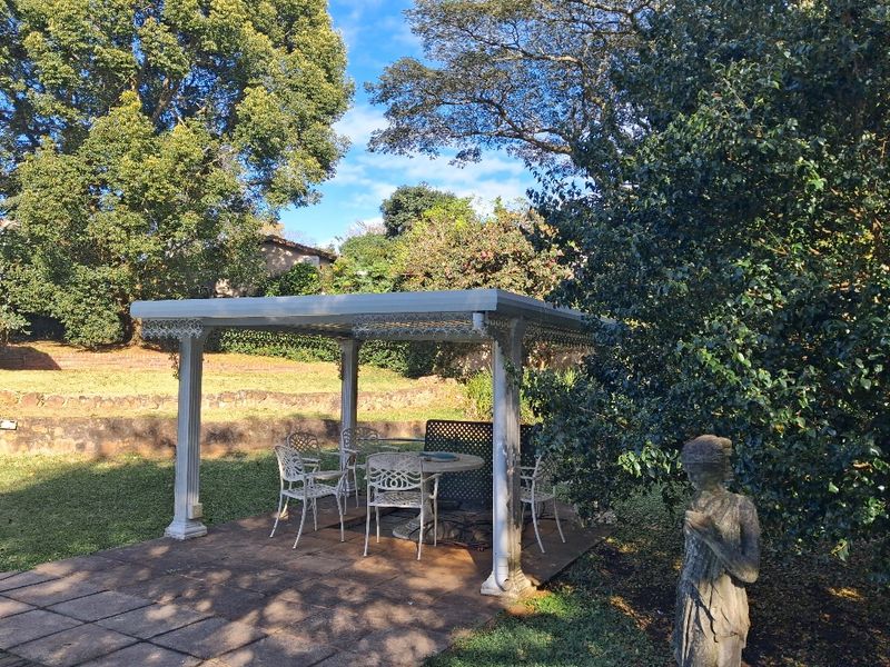 A covered outdoor patio area featuring decorative metal columns and paved flooring, set within a mature garden environment.