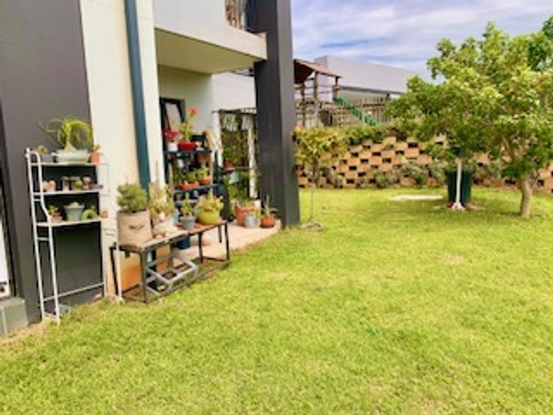 A rear garden view featuring a lawn, a retaining wall, and a paved patio area adjacent to the apartment building.
