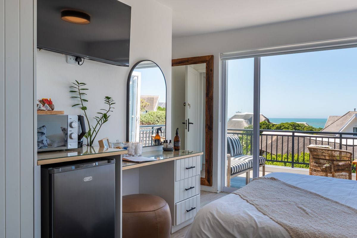 A bright bedroom featuring a built-in vanity, wall-mounted television, and sliding glass doors that open onto a balcony with scenic coastal views.