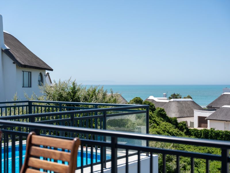A scenic elevated view overlooking the ocean, featuring a modern balcony with glass and metal railings, thatched roof architecture, and a private swimming pool.