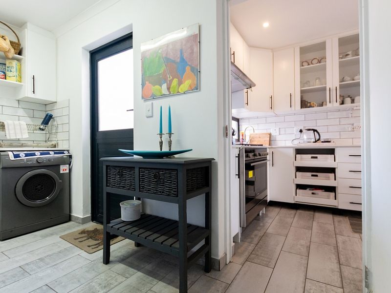 A functional scullery and laundry area featuring white cabinetry, subway tile backsplashes, and modern ceramic flooring, leading into a well-equipped kitchen.