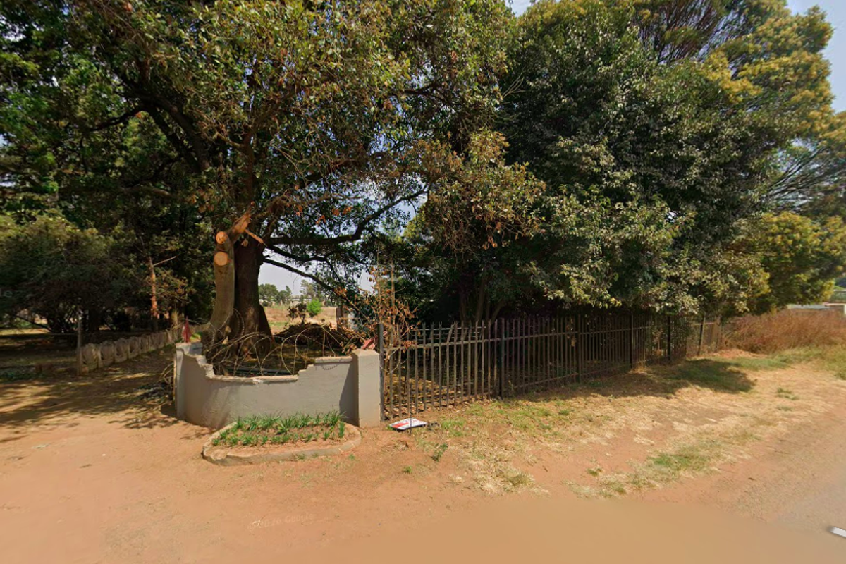 Street view of the property boundary featuring mature trees, metal fencing, and a masonry wall with visible damage.