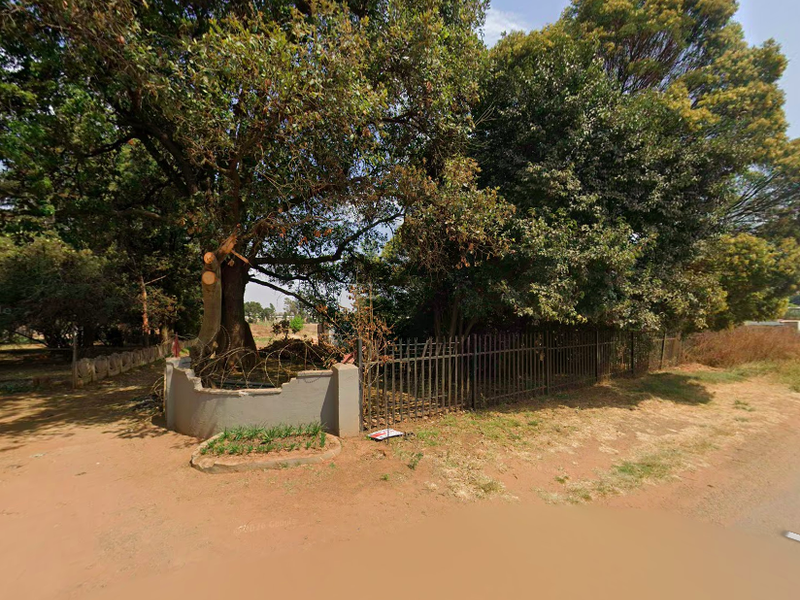 Street view of the property boundary featuring mature trees, metal fencing, and a masonry wall with visible damage.