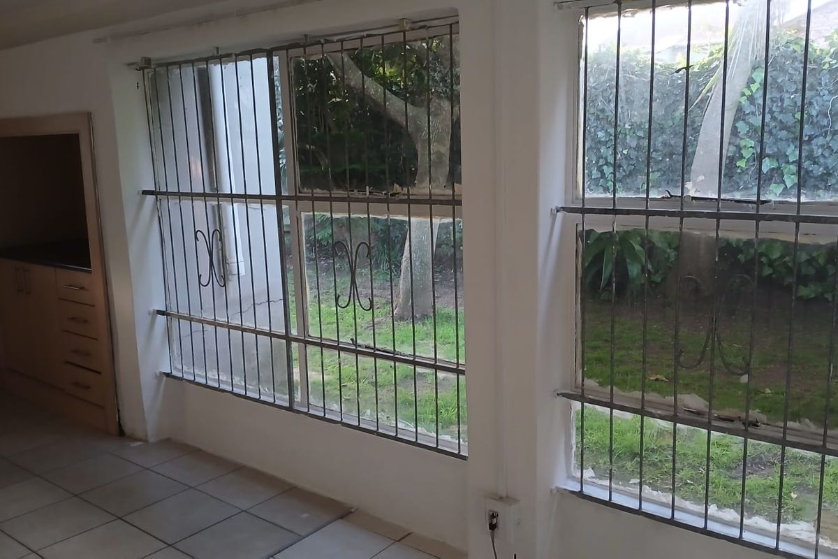 An empty kitchen area featuring tiled flooring, built-in cabinetry, and large windows with security bars overlooking the garden.