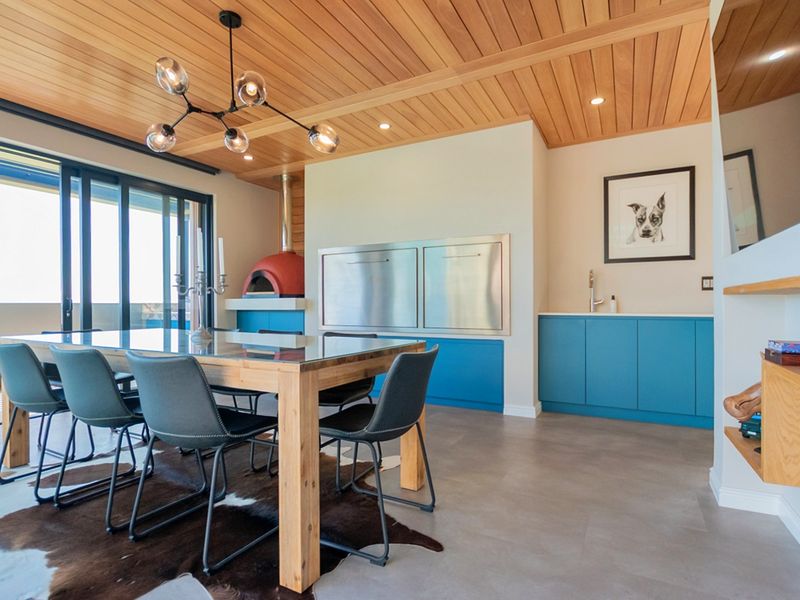 Modern dining area featuring a timber-clad ceiling, built-in pizza oven, stainless steel braai, and custom blue cabinetry with integrated sink.
