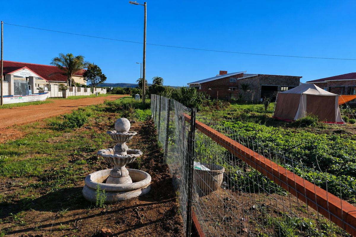 A view of a residential plot featuring a wire mesh fence, a decorative stone fountain, and surrounding neighborhood infrastructure.
