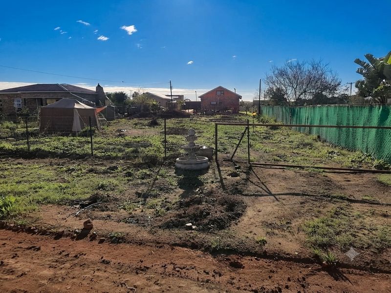 A vacant residential plot featuring a stone fountain, wire fencing, and green shade netting, offering potential for landscaping and development.