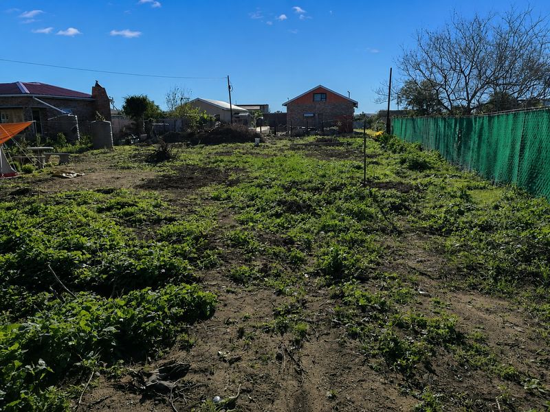 A vacant residential plot featuring overgrown ground cover, perimeter fencing, and a water storage tank, offering a blank canvas for future development.
