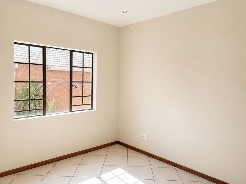 A clean, empty bedroom featuring neutral wall paint, tiled flooring, and a metal-framed window overlooking an exterior brick wall.