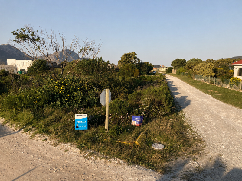 A view of a vacant residential plot situated along a gravel road, featuring natural vegetation and existing utility access points.