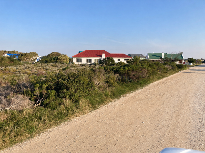A view of a vacant residential plot featuring natural coastal vegetation, adjacent to a gravel road with neighboring houses visible in the background.