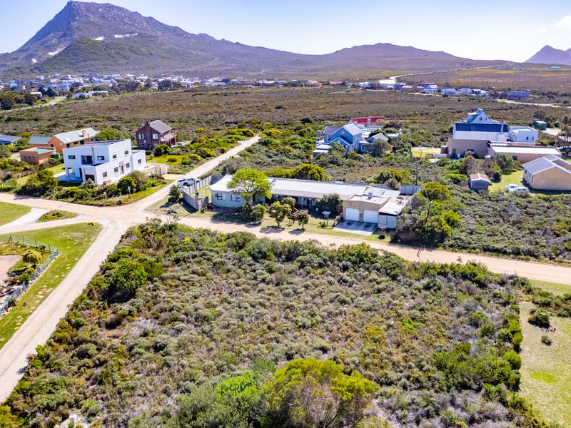 An aerial view of a vacant residential plot featuring natural vegetation, dirt road access, and surrounding neighborhood infrastructure with mountain views.