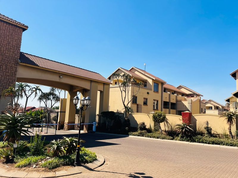 The entrance to the apartment complex features a secure gated access point, paved driveway, and well-maintained perimeter walls with established landscaping.