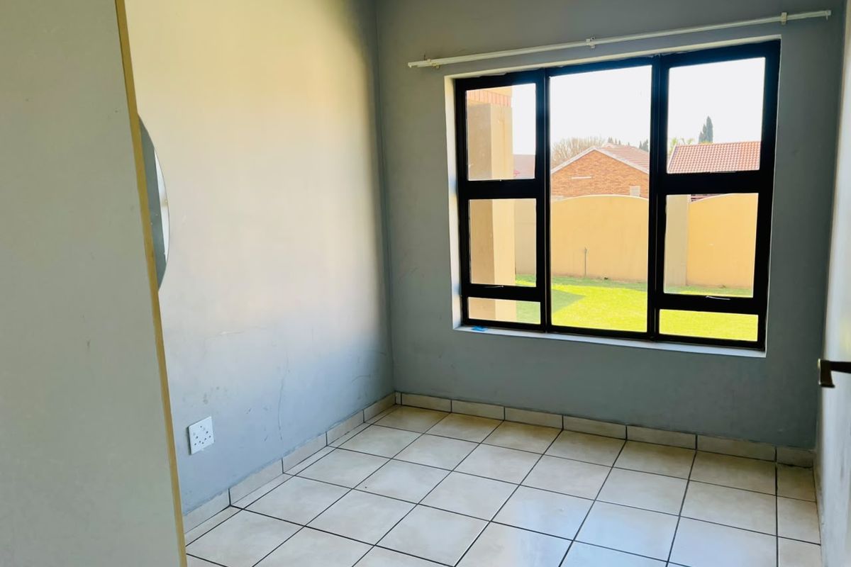 A bright, empty bedroom featuring neutral tiled flooring and large aluminium-framed windows overlooking the garden area.