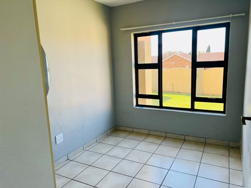 A bright, empty bedroom featuring neutral tiled flooring and large aluminium-framed windows overlooking the garden area.