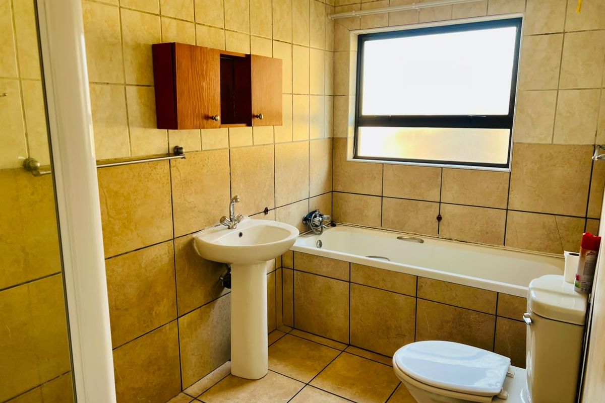 A functional bathroom featuring a pedestal basin, bathtub, toilet, and wall-mounted storage cabinet, finished with neutral wall and floor tiling.