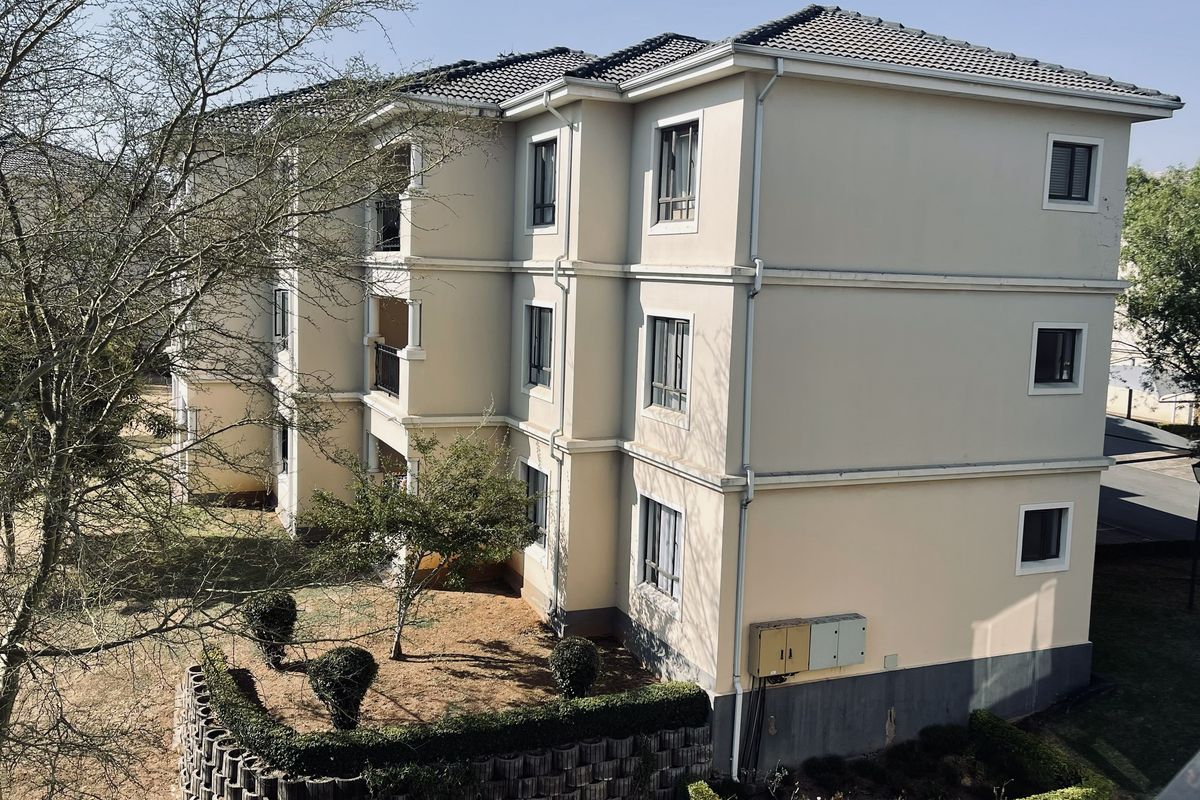 A multi-story apartment building exterior featuring a tiled roof, neutral facade, and integrated balconies overlooking a landscaped garden area with a retaining wall.