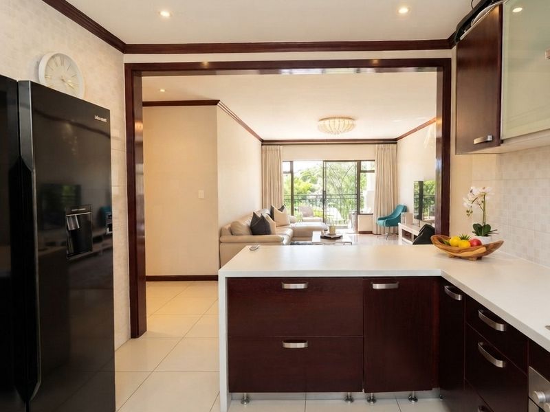 An open-plan kitchen and living area featuring dark wood cabinetry, white stone countertops, and seamless flow to a balcony with natural light.