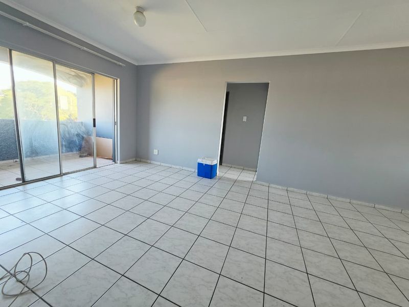 An empty, neutrally painted living room featuring ceramic floor tiles and a large sliding glass door providing access to an outdoor balcony area.