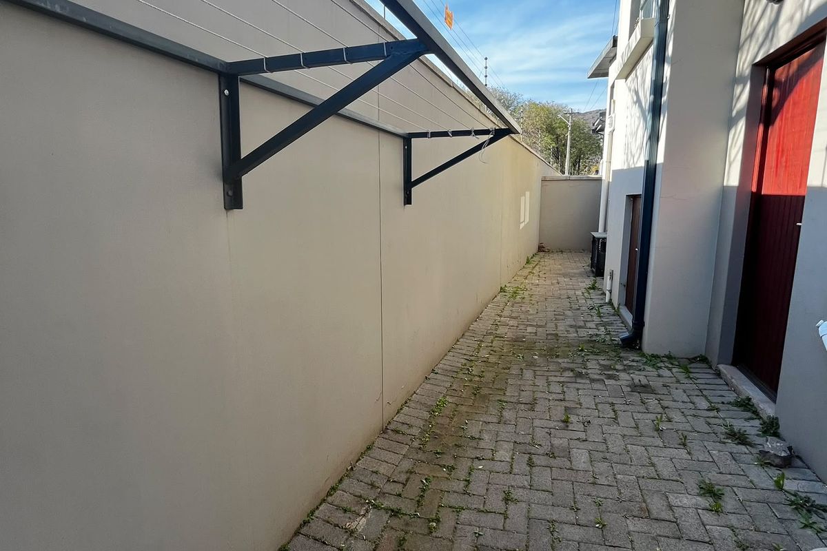 A narrow rear exterior walkway featuring paved flooring, a wall-mounted clothesline, and perimeter walling with electric fencing.
