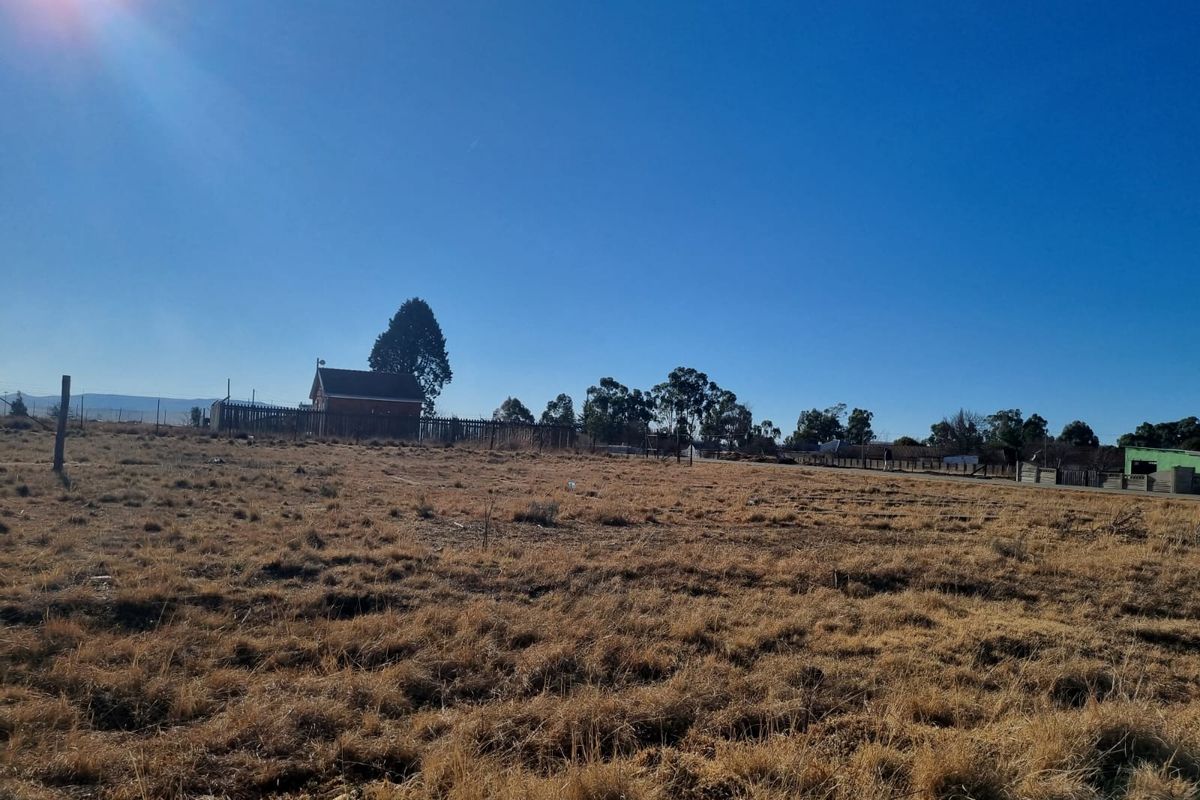 A wide-angle view of a vacant residential plot featuring dry grass, perimeter fencing, and distant structures under a clear blue sky.