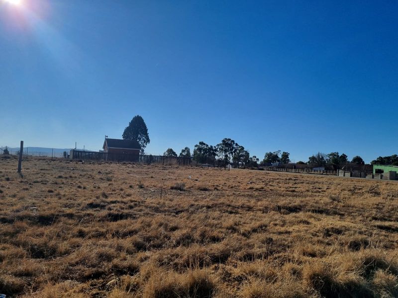 A wide-angle view of a vacant residential plot featuring dry grass, perimeter fencing, and distant structures under a clear blue sky.