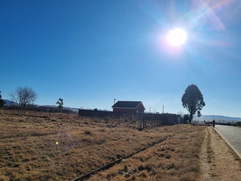 A view of a vacant residential plot featuring dry grass, perimeter fencing, and direct access to a paved road.