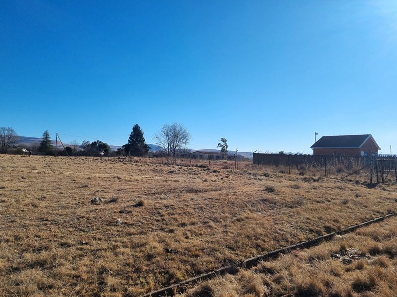 A view of a vacant residential plot featuring dry grass, perimeter wire fencing, and a concrete curb boundary.