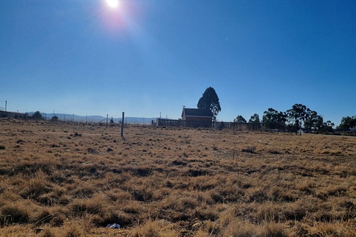 A wide view of a vacant residential plot featuring dry grass and perimeter fencing with a distant structure visible in the background.