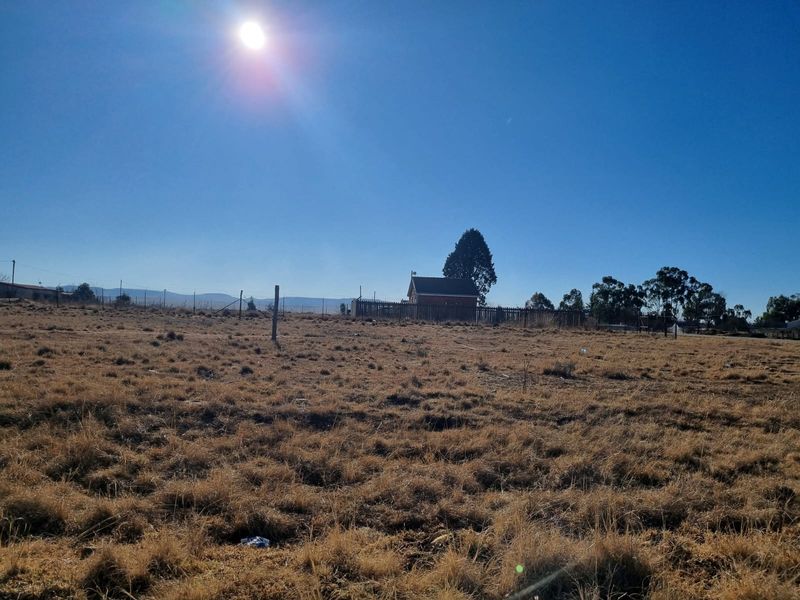 A wide view of a vacant residential plot featuring dry grass and perimeter fencing with a distant structure visible in the background.