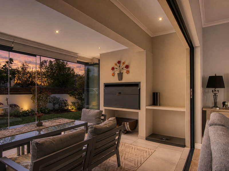 A well-maintained covered patio featuring a built-in fireplace, recessed lighting, and seamless stacking glass doors that open to a landscaped garden.