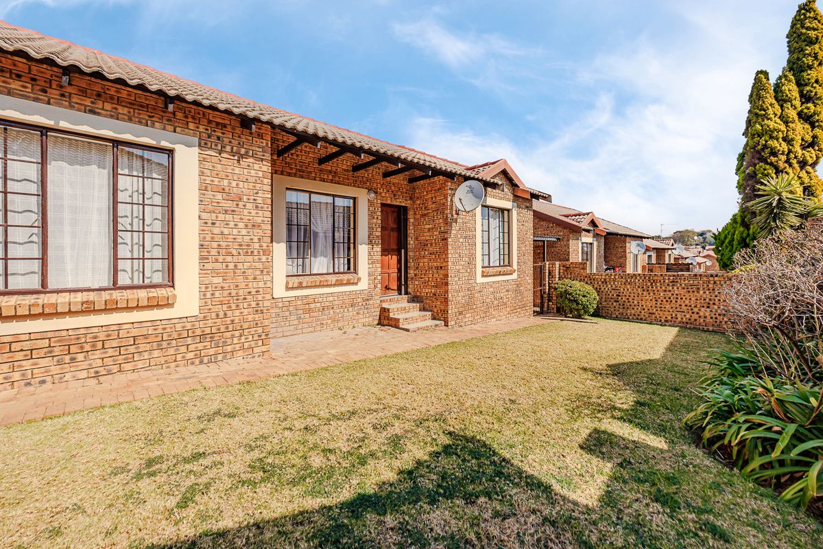 A well-maintained townhouse exterior featuring a face-brick facade, tiled roof, paved patio area, and a private lawn with boundary walling.