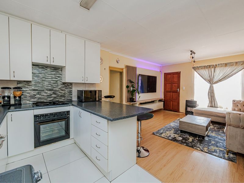 Open-plan living area featuring a modern kitchen with built-in oven, hob, and mosaic backsplash, flowing into a comfortable lounge space with laminate flooring.