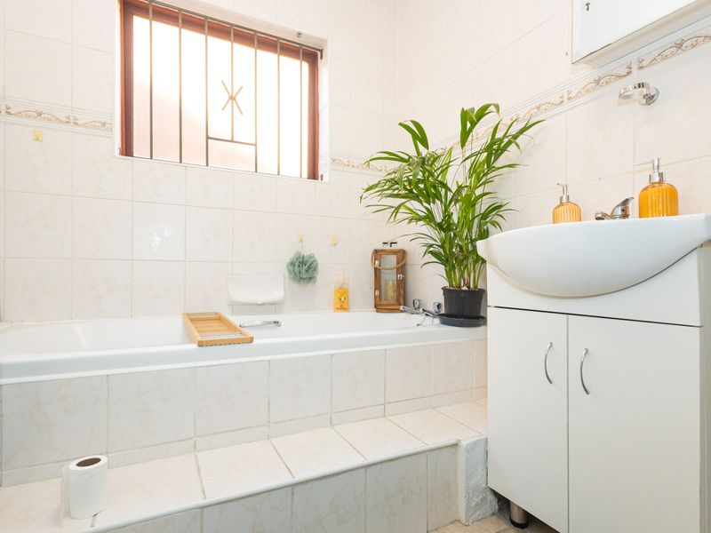 A functional bathroom featuring a built-in bathtub, vanity cabinet, and tiled finishes, with a window fitted with security bars.