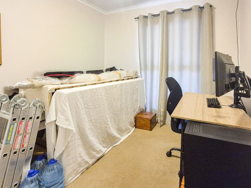 A functional bedroom space featuring neutral wall finishes, carpet flooring, and window treatments, currently utilized as a home office and storage area.