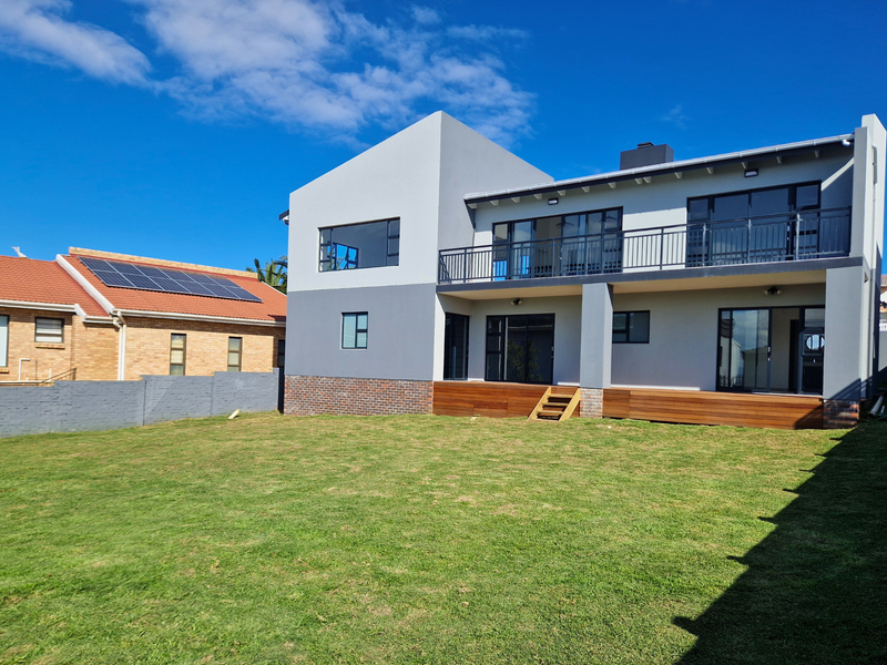 Rear view of a modern two-story house featuring a spacious lawn, timber decking, and a large upper-level balcony with sliding glass doors.