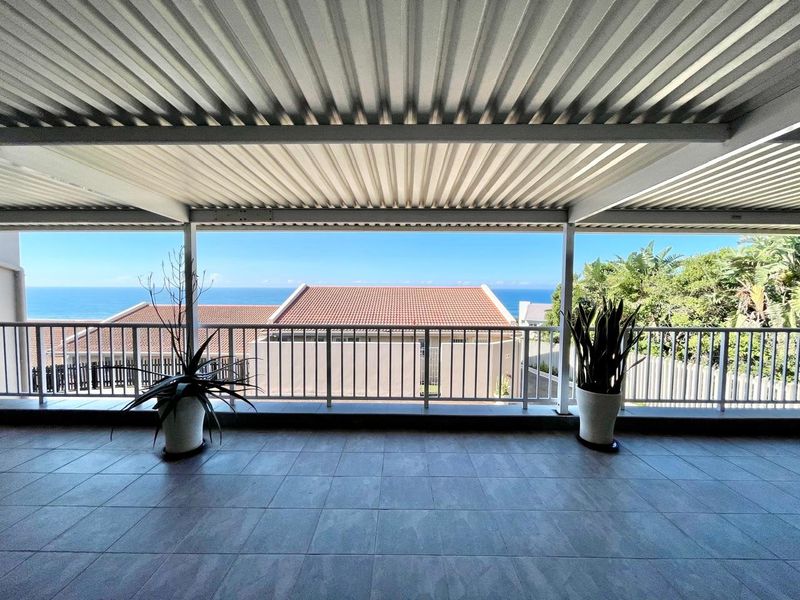 A spacious covered balcony featuring tiled flooring, a corrugated metal roof, and a metal balustrade, offering clear views of the ocean and surrounding neighborhood.