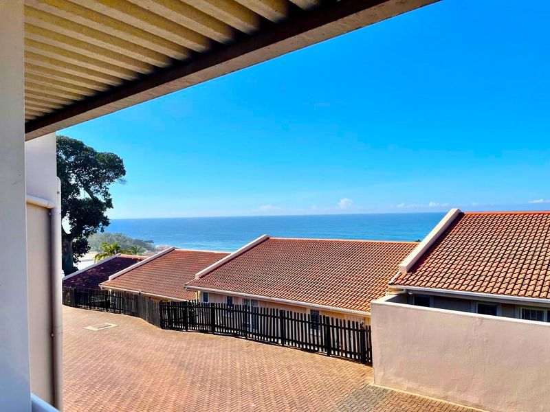 An elevated view overlooking a residential complex with tiled roofs, a paved driveway, and a clear ocean horizon in the background.