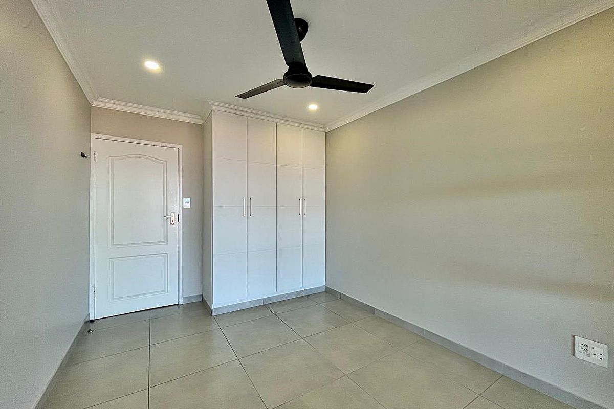 A clean, empty bedroom featuring neutral tiled flooring, built-in wardrobes, a modern ceiling fan, and recessed lighting.