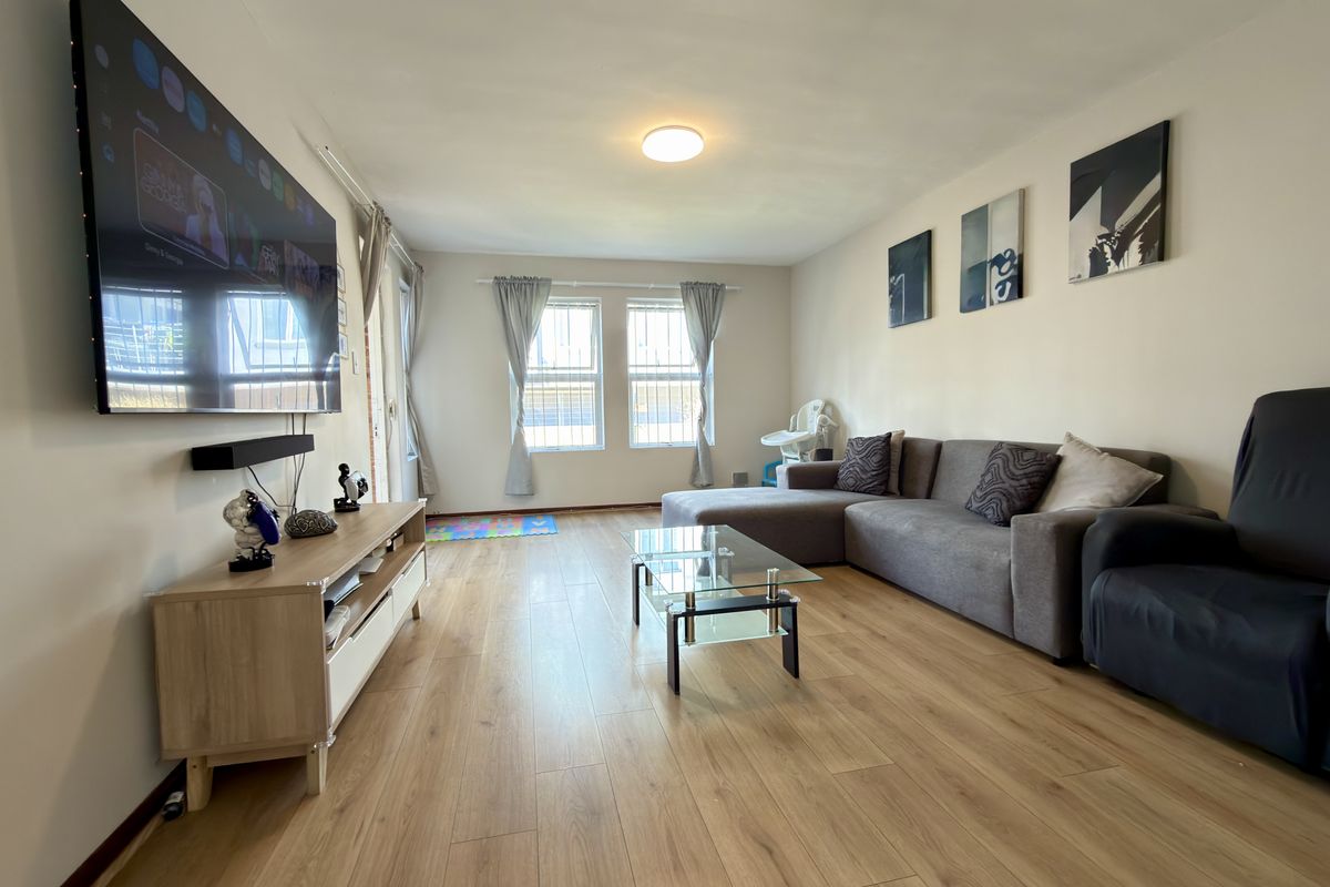 A bright and spacious living room featuring light wood-look laminate flooring and large windows fitted with security bars.