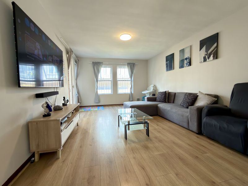 A bright and spacious living room featuring light wood-look laminate flooring and large windows fitted with security bars.