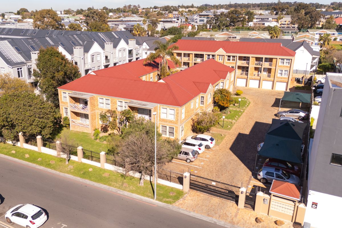 Aerial view of a multi-story apartment complex featuring facebrick construction, red metal roofing, paved parking areas, and established garden surroundings.