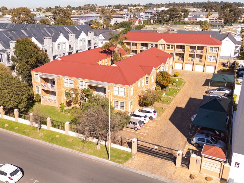 Aerial view of a multi-story apartment complex featuring facebrick construction, red metal roofing, paved parking areas, and established garden surroundings.